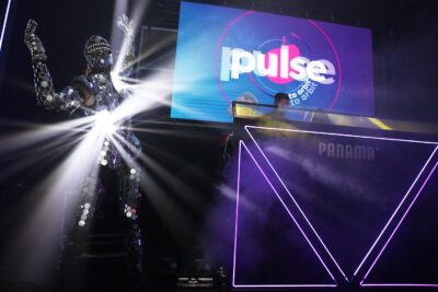 A performer in a reflective, mirrored outfit raises their arms on a dimly lit stage, embodying the essence of AI Wonderland. Bright lights shine from their suit as a DJ booth with neon accents and a screen displaying the logo "Pulse" graces the background, capturing the futuristic vibe of Customer Success.