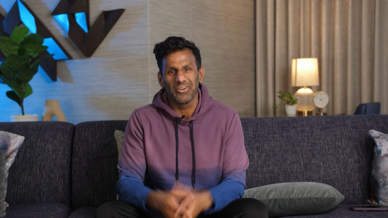 Nick Mehta is wearing wearing a purple hoodie sitting on a dark sofa in a modern living room. Behind him, a lamp, clock, and plant rest on a table. The wall features geometric decor and curtains, creating a cozy atmosphere.