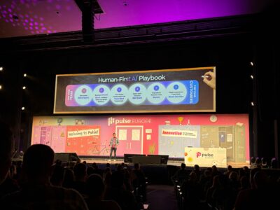 At Pulse Europe 2024, a speaker stands onstage in front of a large screen displaying the "Human-First AI Playbook," heralding the future of Customer Success. The colorful stage is adorned with vibrant signs and illustrations, while the engaged audience is visible in the foreground.