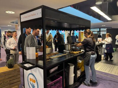 A woman prepares stroopwafels at the "Fresh Dutch Waffles" stall, drawing a crowd eager for a taste. The modern indoor setting, with its sleek furnishings and overhead lighting, buzzes like Pulse Europe 2024's lively atmosphere focused on customer success.