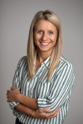 A person with long blonde hair smiles at the camera, wearing a green and white striped shirt with sleeves rolled up. Their arms are confidently crossed in front, exuding an air of empowerment fitting for a Women In CS Masterclass Series. The background is a plain, neutral gray.