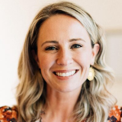 A smiling woman with long, wavy blonde hair, wearing large circular earrings and a light-colored top, stands against a soft-focus background. She radiates cheerfulness and approachability, embodying the PowerUp spirit of the Women In CS Masterclass Series.