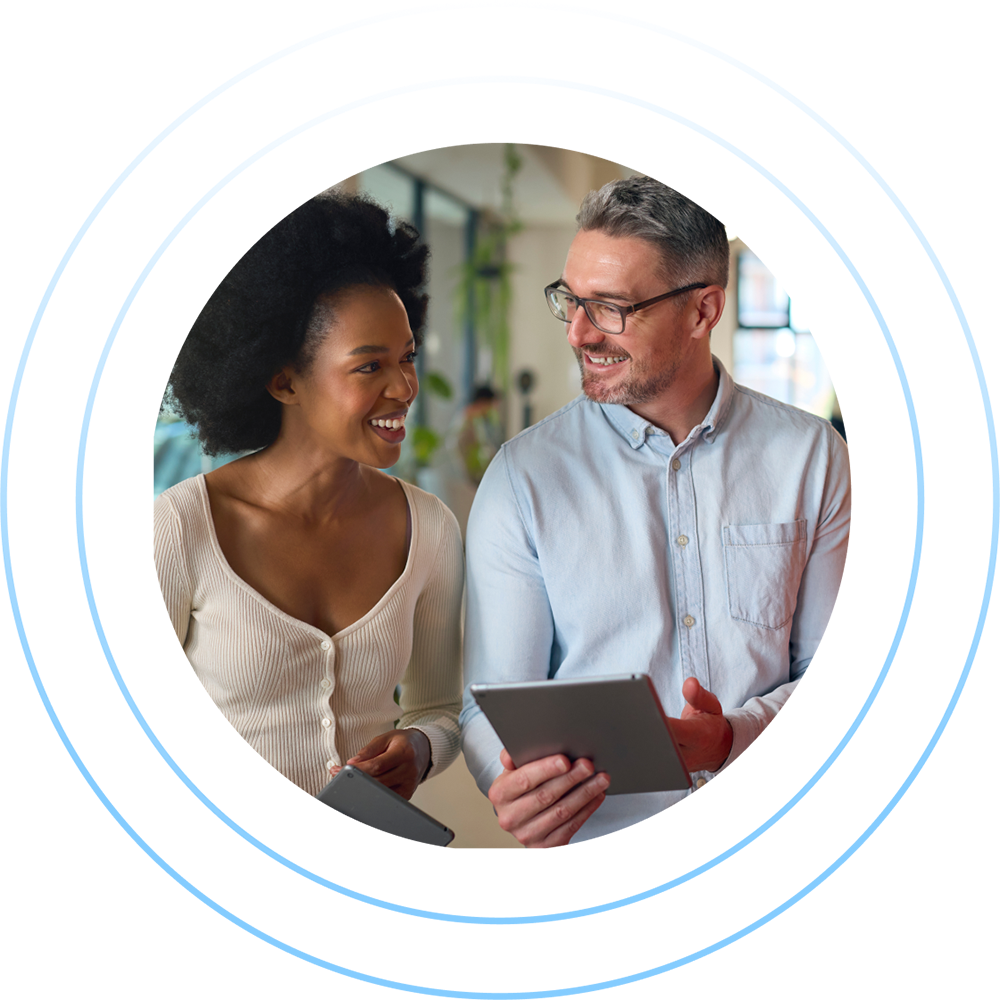 A man and woman are smiling and holding tablets while standing in an office. The woman, wearing a white top, and the man, in a light blue shirt, seem engaged in a pleasant conversation about the latest Customer Success Index.