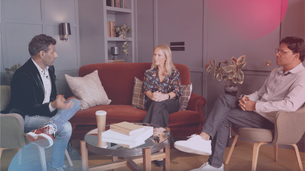 Three people are sitting in a modern living room with a red sofa and plants. A man in a black jacket and jeans excitedly discusses the Gainsight event with a woman in a floral blouse and another man in a white shirt. Coffee cups and books are on the glass table.